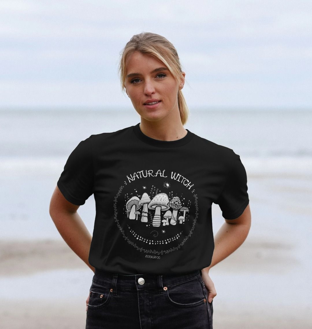 Blonde haired model outside on the Isle of Wight, wearing a black unisex t-shirt with white illustrations of mushrooms, toadstools, moons, stars, leaves and vines, with the words Natural Witch in funky typography, halloween spooky design by MooksGoo