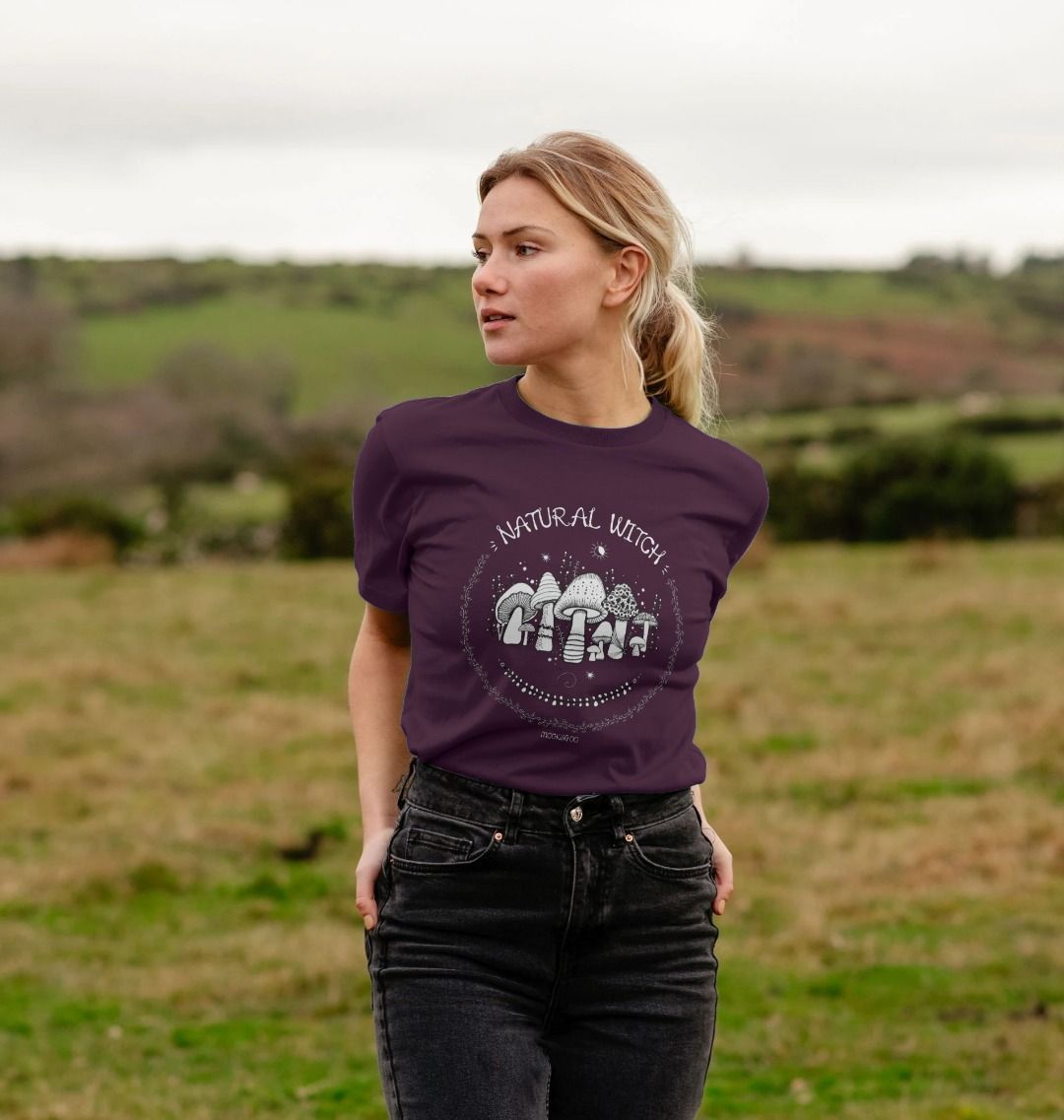 Blonde haired model outside on the Isle of Wight, wearing a dark purple unisex t-shirt with white illustrations of mushrooms, toadstools, moons, stars, leaves and vines, with the words Natural Witch in funky typography, halloween spooky design by MooksGoo