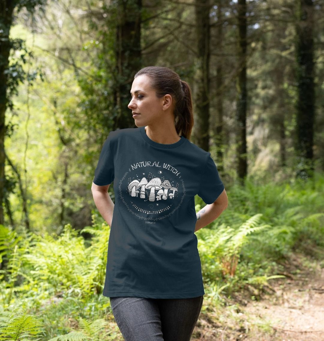 Blonde haired model outside on the Isle of Wight, wearing a denim blue unisex t-shirt with white illustrations of mushrooms, toadstools, moons, stars, leaves and vines, with the words Natural Witch in funky typography, halloween spooky design by MooksGoo