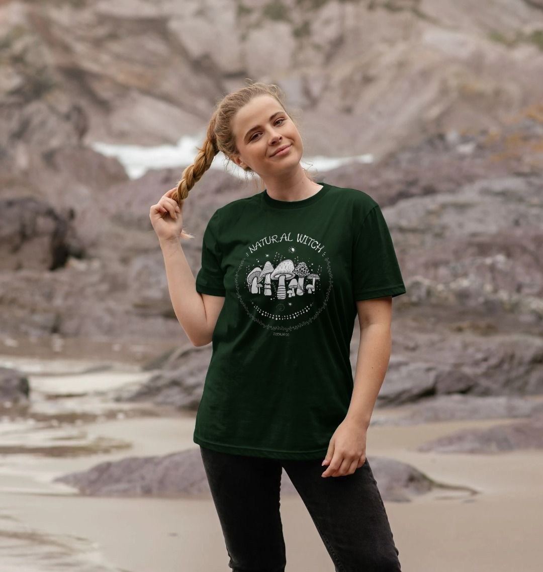Blonde haired model outside on the Isle of Wight, wearing a evergreen unisex t-shirt with white illustrations of mushrooms, toadstools, moons, stars, leaves and vines, with the words Natural Witch in funky typography, halloween spooky design by MooksGoo