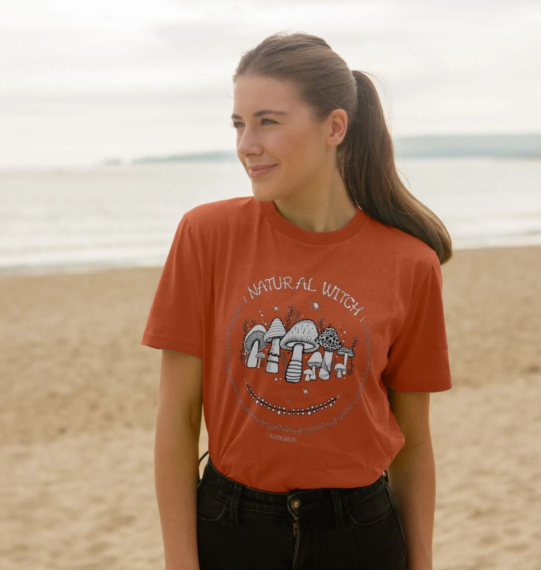 Blonde haired model outside on the Isle of Wight, wearing a rusty orange unisex t-shirt with white illustrations of mushrooms, toadstools, moons, stars, leaves and vines, with the words Natural Witch in funky typography, halloween spooky design by MooksGoo