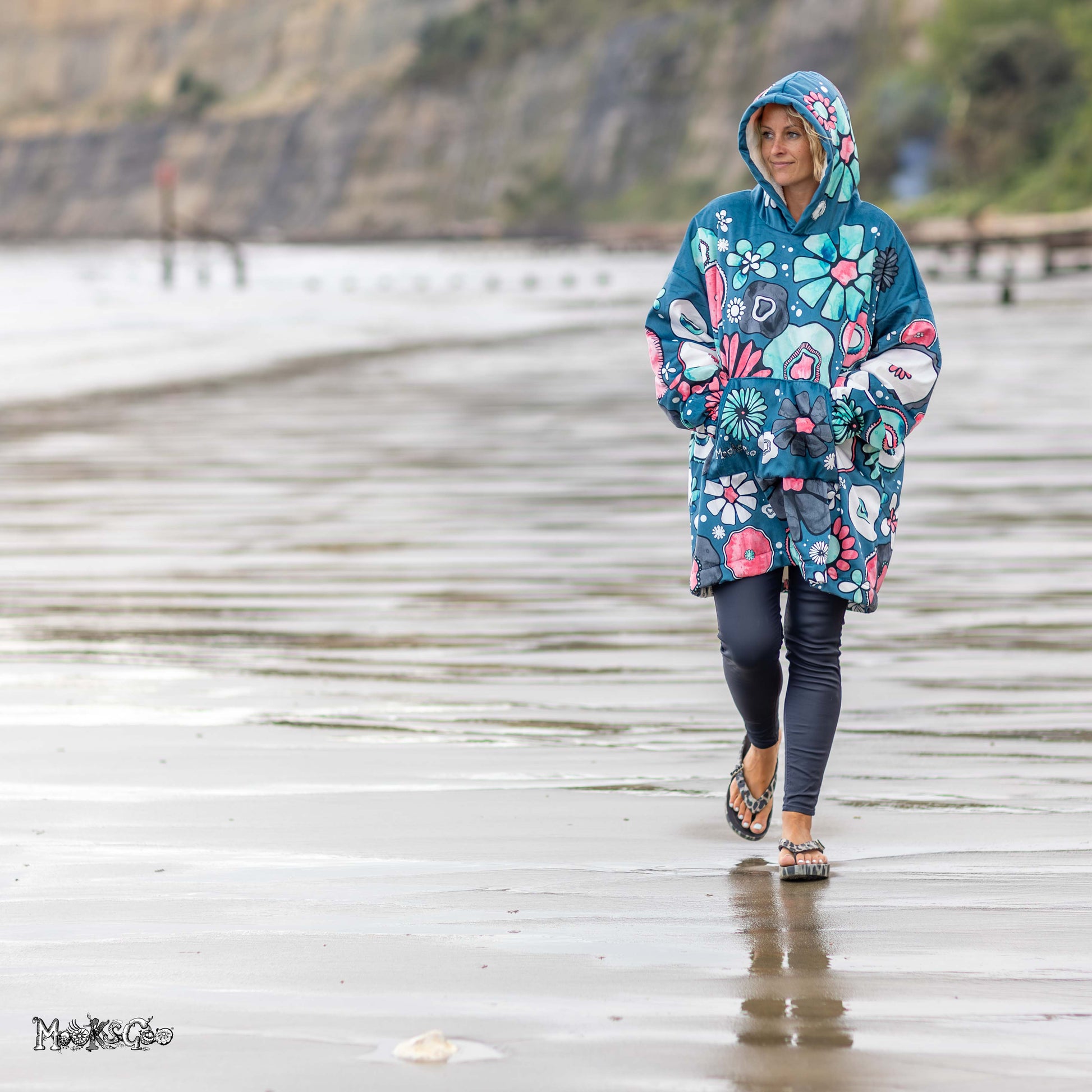 A woman walks on a wet sandy beach wearing a teal hooded poncho with a bold and colourful floral print, along with dark leggings and sandals. The background shows a blurred beach and cliffs. Hoodie designed by MooksGoo