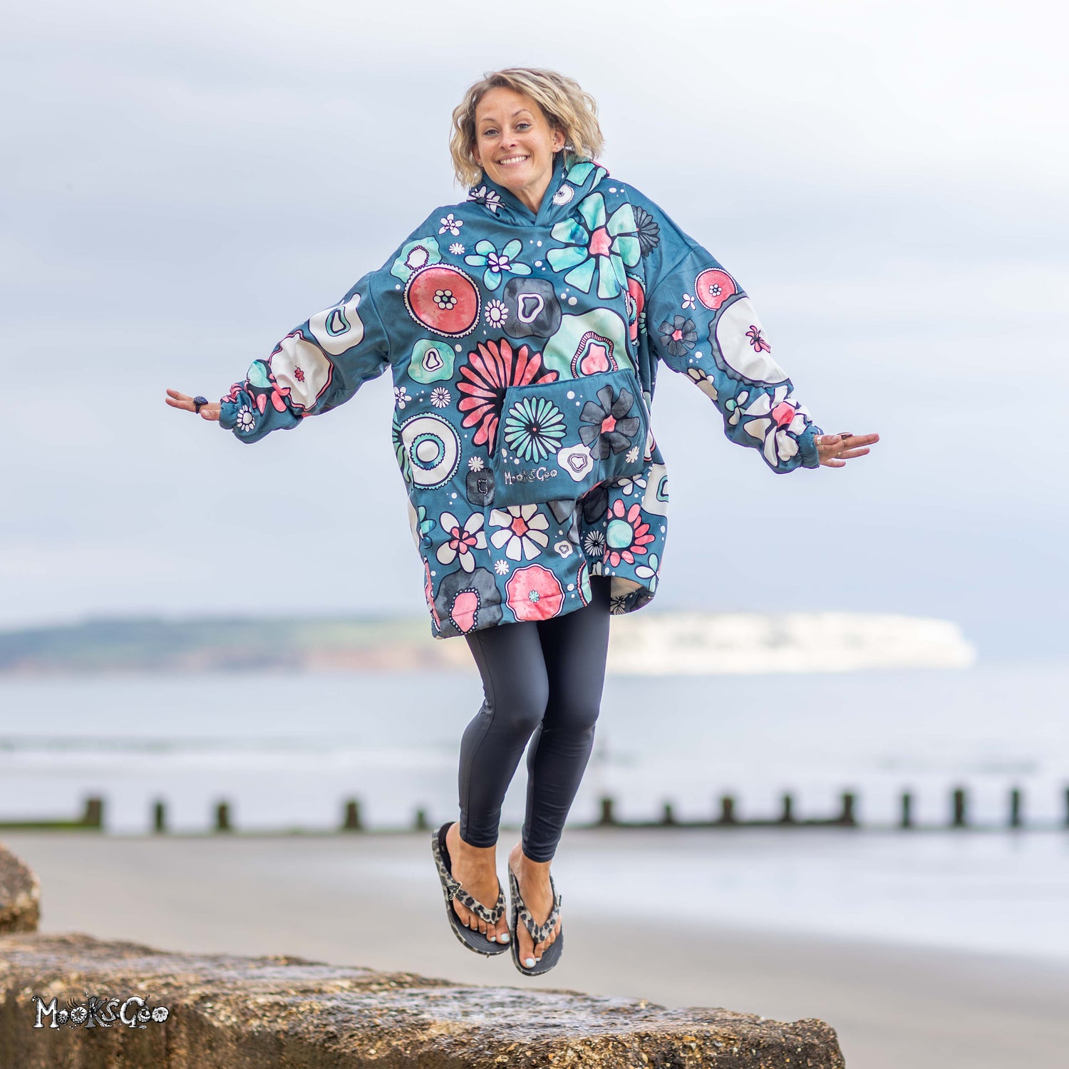 A woman jumps on a beach wearing an oversized teal blue hooded jacket covered in colourful floral and circular patterns, with black leggings and slip-on shoes. Designed by MooksGoo