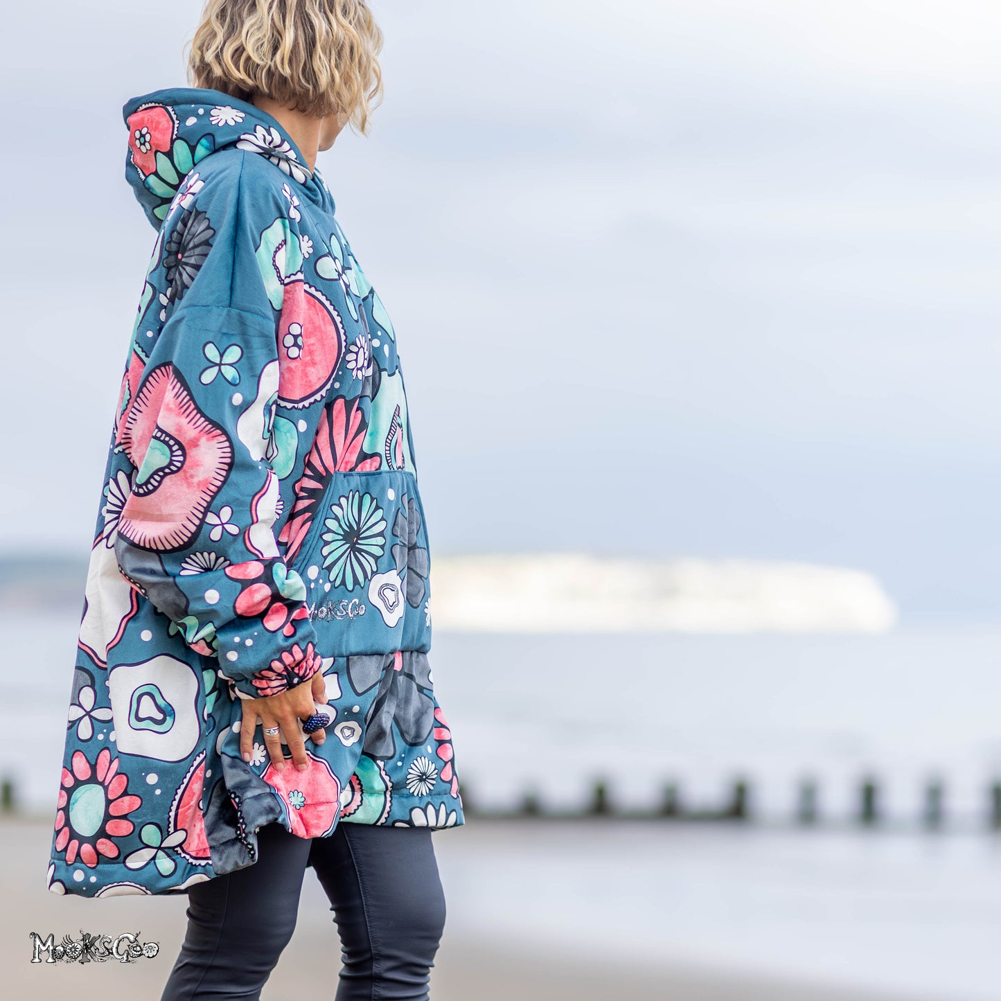 A woman stands on a beach wearing a bold, colourful hooded jacket hoodie with illustrated floral print in teal, pink, white, and black. The background shows a soft-focus view of the sea and sky. Designed by MooksGoo
