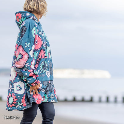 A woman stands on a beach wearing a bold, colourful hooded jacket hoodie with illustrated floral print in teal, pink, white, and black. The background shows a soft-focus view of the sea and sky. Designed by MooksGoo