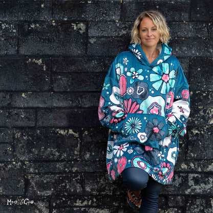 A woman wearing a teal blue hooded blanket jacket hoodie with a colourful illustrated floral pattern, posing against a dark textured brick wall. Hoodie designed by MooksGoo