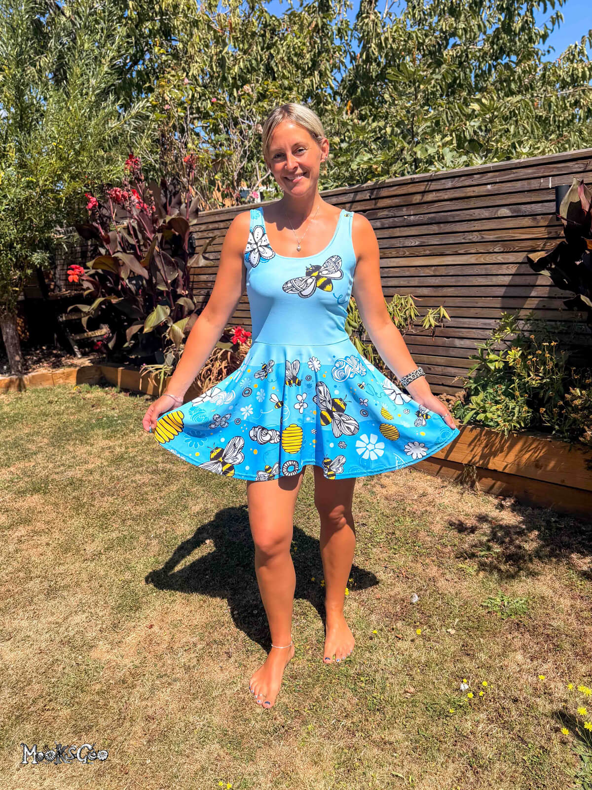 A woman wearing a light blue sleeveless flared short dress with a bees, honey and floral print, standing barefoot on a grassy garden. Dress designed by MooksGoo