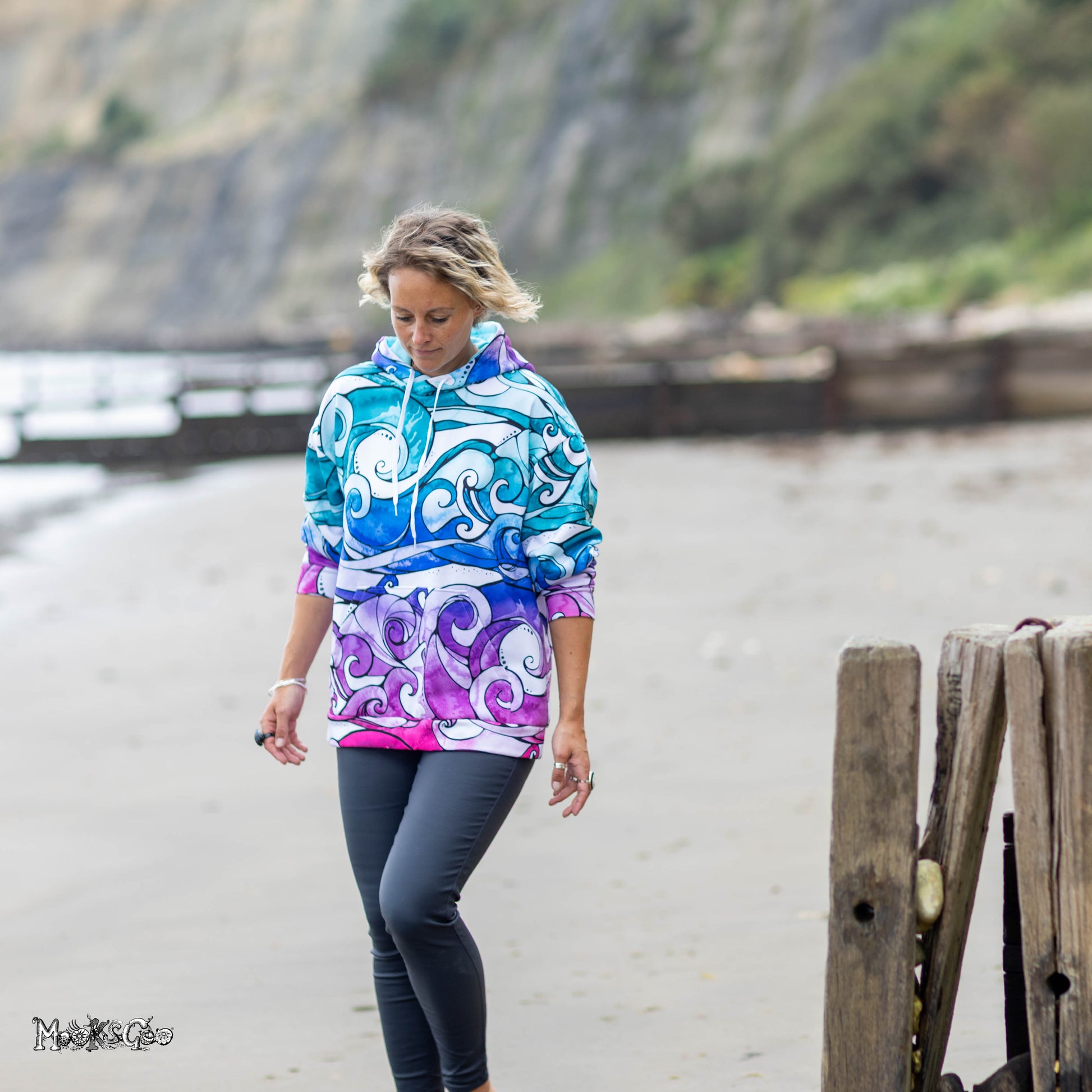 A woman walking on a sandy beach wearing a colourful hoodie with a blue, purple, and white wave pattern, paired with dark grey leggings. The background shows a blurred coastal landscape with cliffs and water. Hoodie designed by MooksGoo