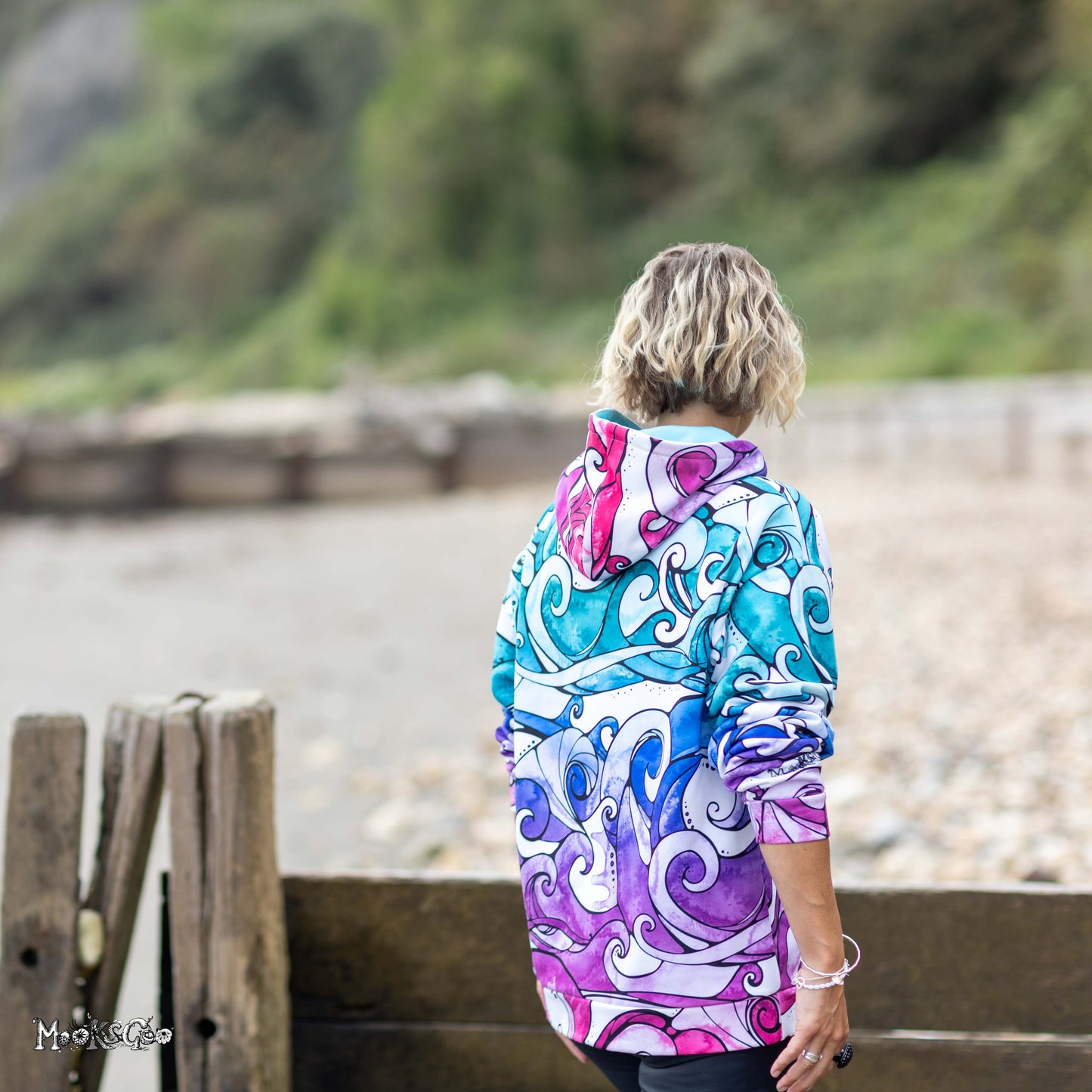 A woman viewed from behind wearing a colourful rainbow coloured hooded hoodie with a swirling illustrated wave print in teal, blue, purple, pink, and white, standing outdoors near a wooden barrier with a blurred natural background. Designed by MooksGoo