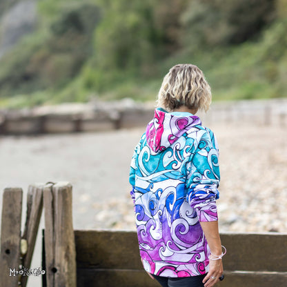A woman viewed from behind wearing a colourful rainbow coloured hooded hoodie with a swirling illustrated wave print in teal, blue, purple, pink, and white, standing outdoors near a wooden barrier with a blurred natural background. Designed by MooksGoo