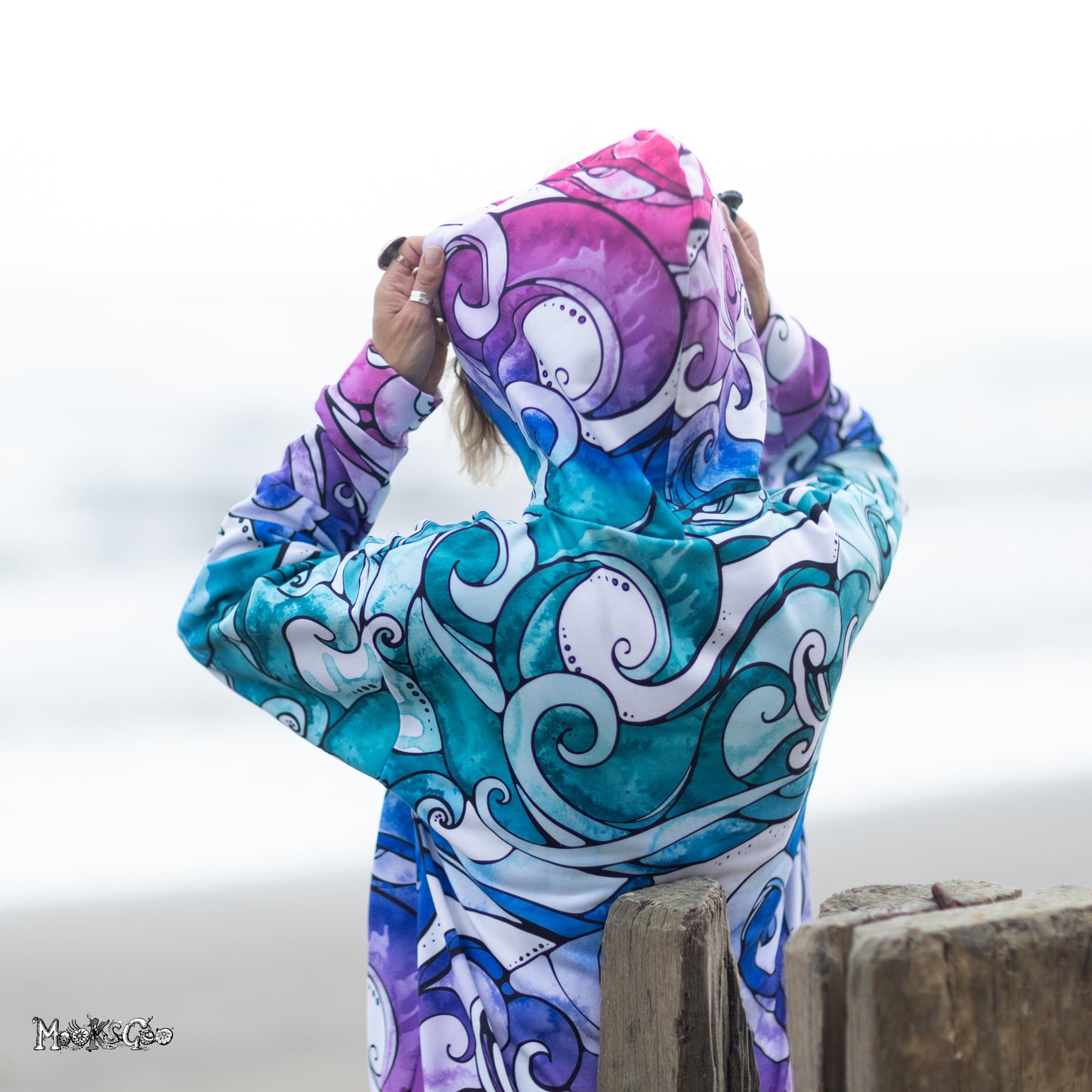 A person viewed from behind wearing a rainbow-coloured long-sleeve hooded top with an abstract surfing wave print in pink, purple, teal, blue, and white, standing outdoors near a wooden post with a blurred light grey background. Hoodie designed by MooksGoo