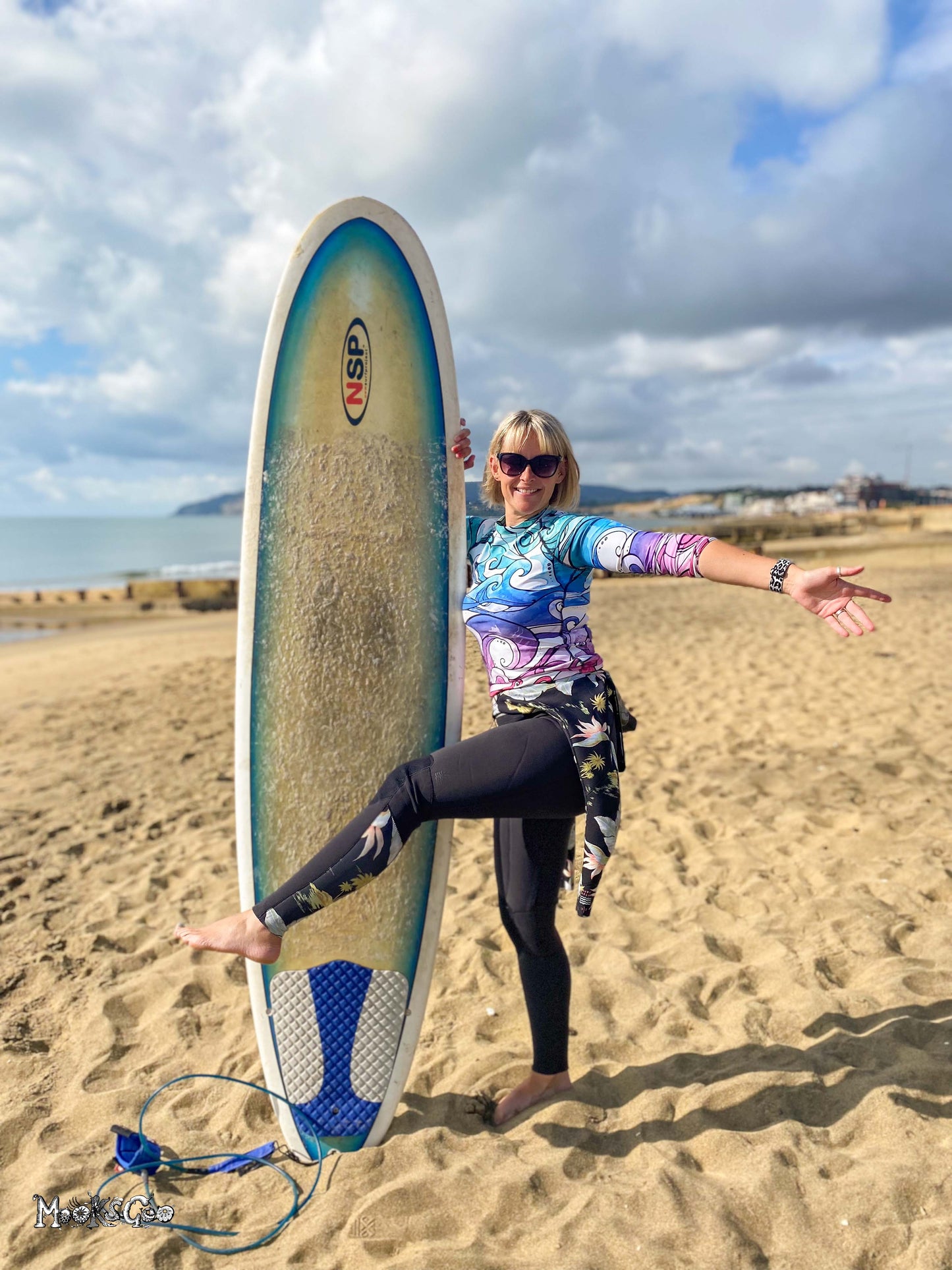 A woman posing on a sandy beach holding a long surfboard upright. She is wearing a colorful patterned long-sleeve rash guard, black leggings, and sunglasses, with the ocean and cloudy sky in the background. Designed by MooksGoo