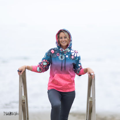 A woman stands on wooden beach stairs wearing a colourful gradient hoodie with an illustrated floral and daisy pattern, dark leggings, and smiling against a misty beach background. Hoodie designed by MooksGoo