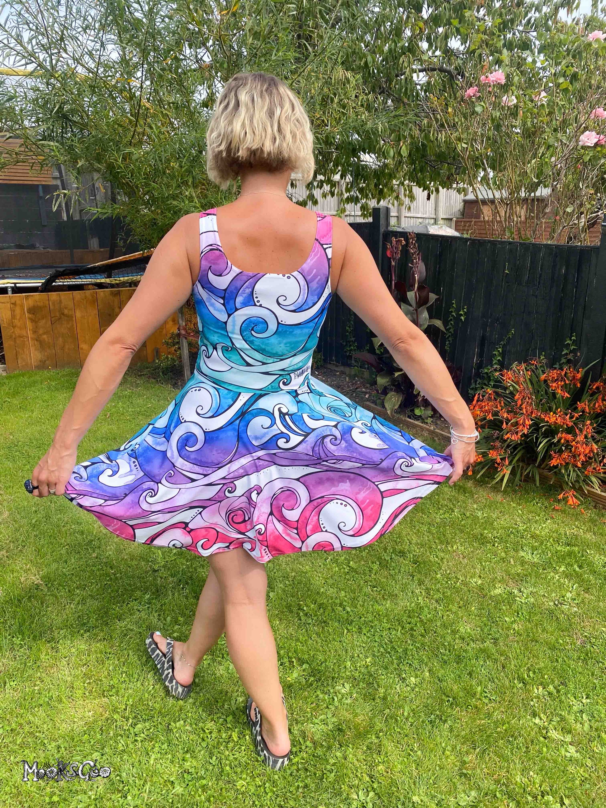 A woman wearing a sleeveless A-line dress with a vibrant blue, purple, and pink swirly wave pattern, photographed from behind in a garden setting. Funky dress designed by MooksGoo