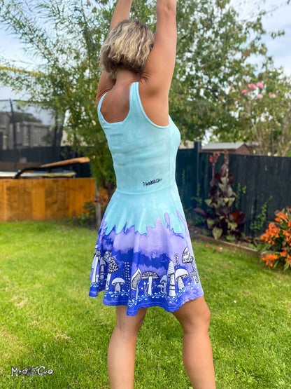 A woman standing outdoors in a backyard wearing a sleeveless skater dress with a light blue and purple gradient and a white mushroom illustration on the skirt. She is seen from behind with her arms raised. Dress designed by MooksGoo