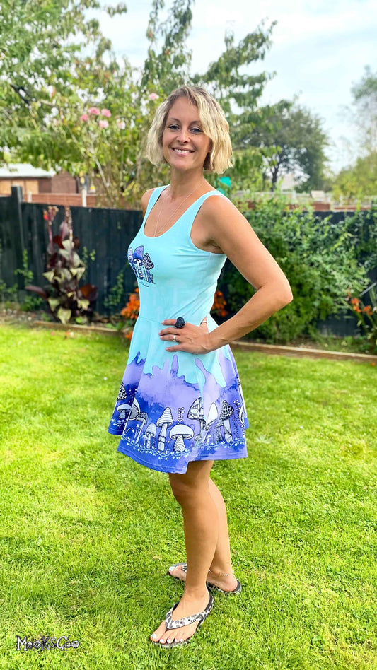 A woman wearing a quirky blue and purple  dress with illustrated mushrooms, standing on green grass. Skater dress designed by MooksGoo