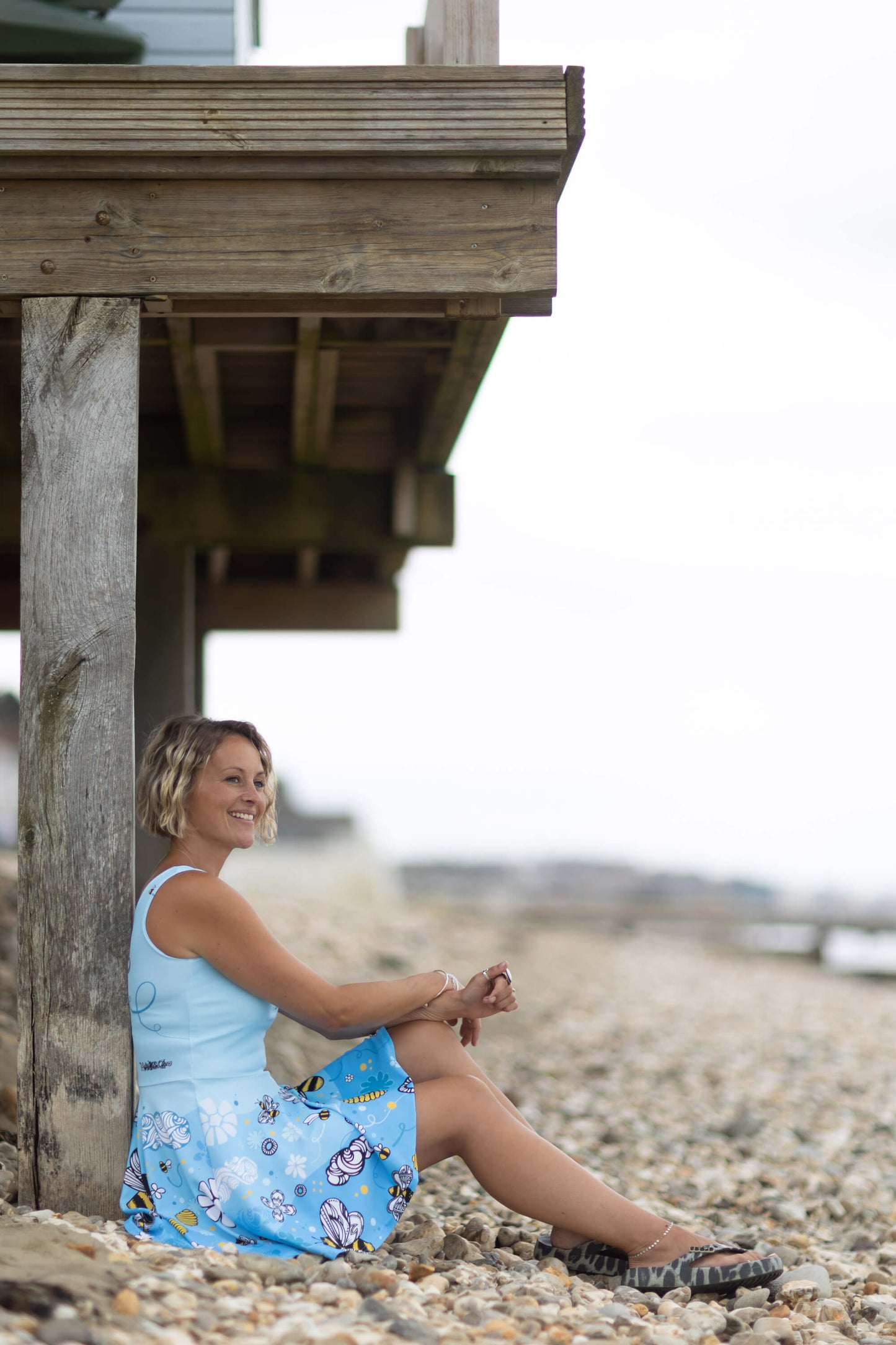 A lady sitting on a pebbly beach wearing a bee and floral short dress, designed by MooksGoo