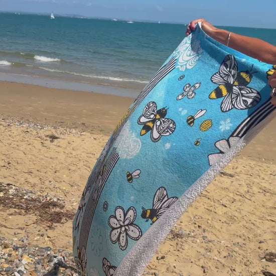 A lady holding a soft blue towel on the beach, with illustrated bees, honey, stripes and flowers, designed by MooksGoo
