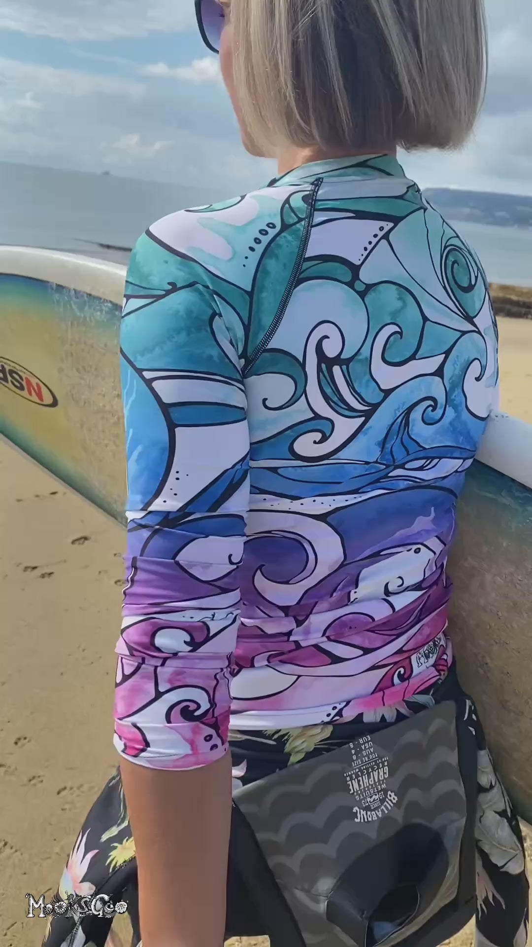 A lady with a surf board wearing a bright and colourful rainbow long sleeved rash guard with swirling waves, designed by MooksGoo