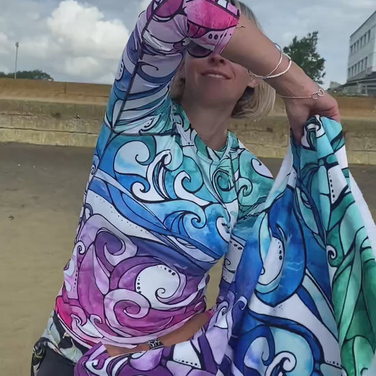 a woman wearing a rainbow wave rash vest is holding a bright funk colourful rainbow-coloured towel with illustrated waves, designed by MooksGoo