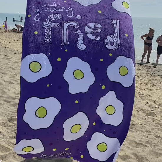 A lady holding a purple towel with illustrated fried eggs and funky typography saying Getting Fried, designed by MooksGoo