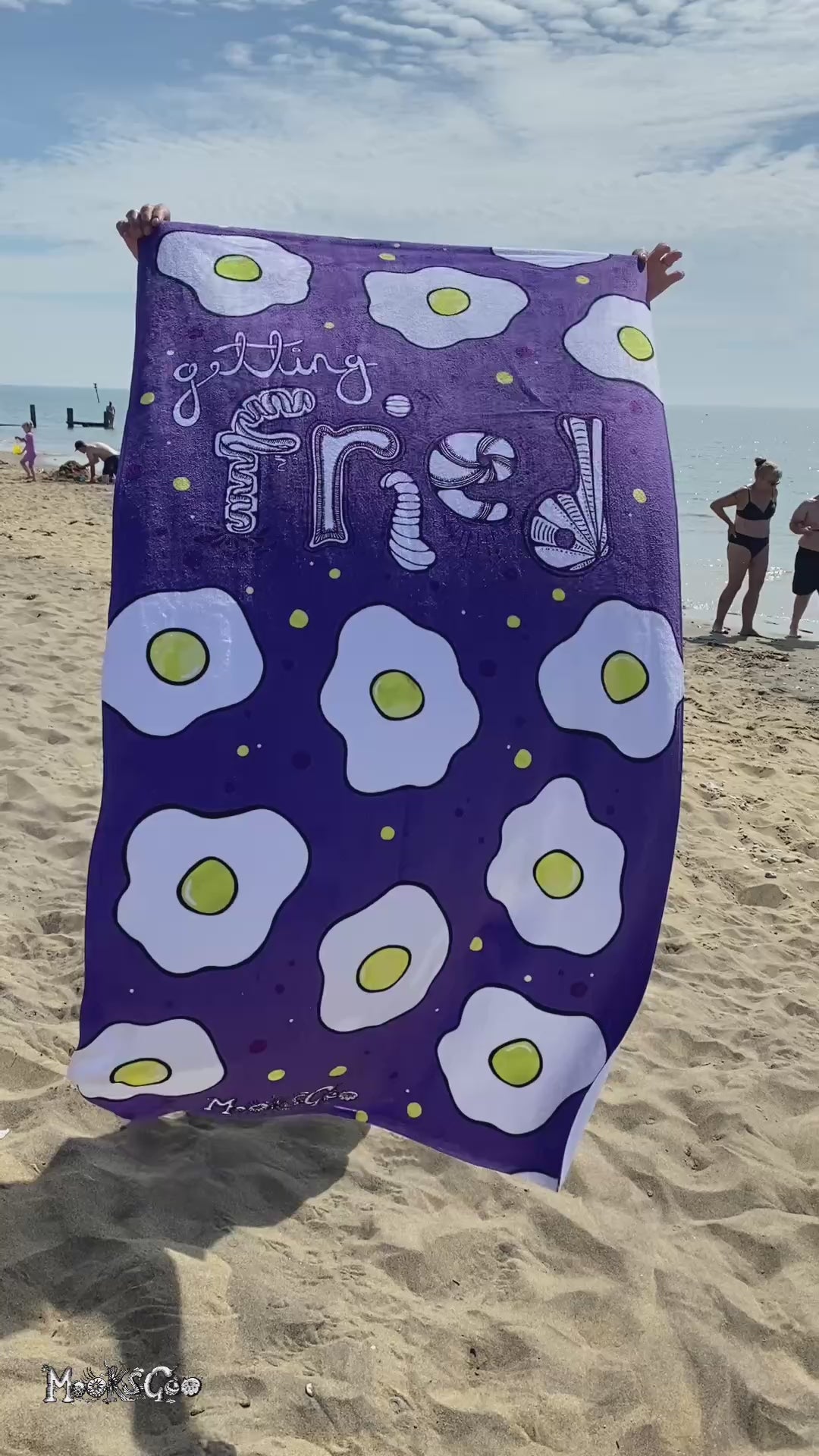 A lady holding a purple towel with illustrated fried eggs and funky typography saying Getting Fried, designed by MooksGoo