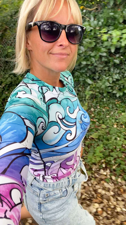 A woman wearing a colourful long-sleeve top with a blue, purple, and white wave pattern, paired with light-wash denim jeans and black sunglasses, photographed outdoors against a background of green foliage. Rash guard designed by MooksGoo