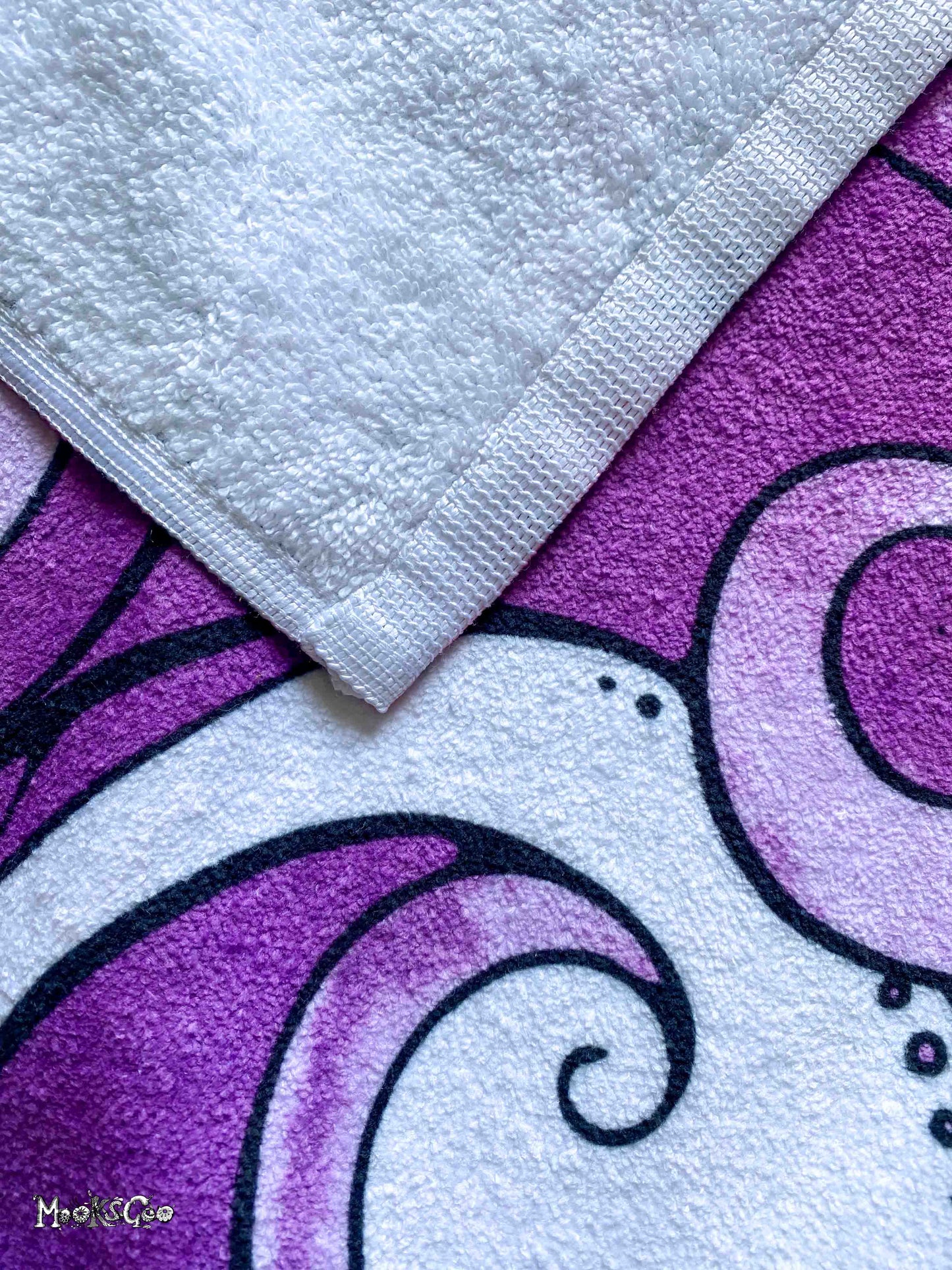 A close-up of a white terry cloth towel with a stitched border laid diagonally over a purple and white patterned beach towel featuring black and white illustrated waves, designed by MooksGoo