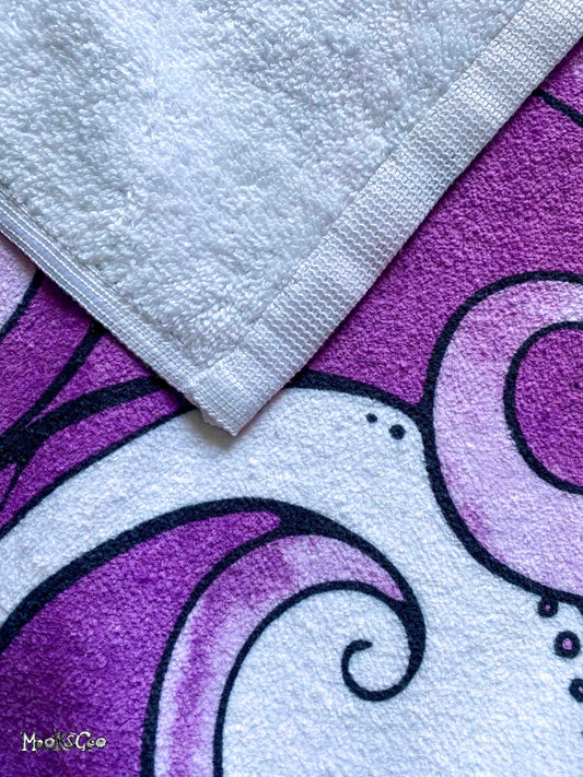 A close-up of a white terry cloth towel with a stitched border laid diagonally over a purple and white patterned beach towel featuring black and white illustrated waves, designed by MooksGoo