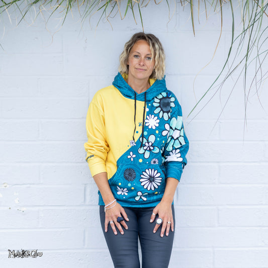 A woman wearing a color-blocked hoodie with a yellow left side and a teal right side decorated with white and black floral illustrated patterns, designed by MooksGoo, standing against a white brick wall.