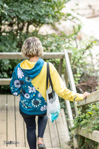A woman viewed from behind walking up wooden steps outdoors, wearing a hooded sweatshirt with a split yellow and teal blue and a white floral print, designed by MooksGoo, while carrying a tote bag.