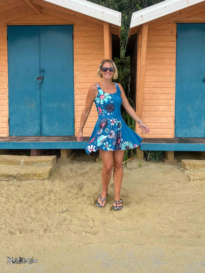 A woman wearing a blue floral sun dress and sunglasses stands on a sandy beach in front of two wooden beach huts with blue doors. Dress designed by MooksGoo