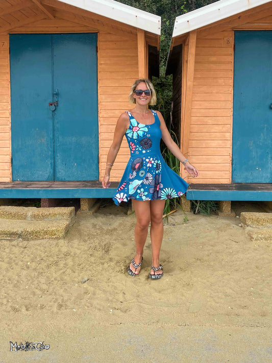 A woman wearing a blue floral sun dress and sunglasses stands on a sandy beach in front of two wooden beach huts with blue doors. Dress designed by MooksGoo